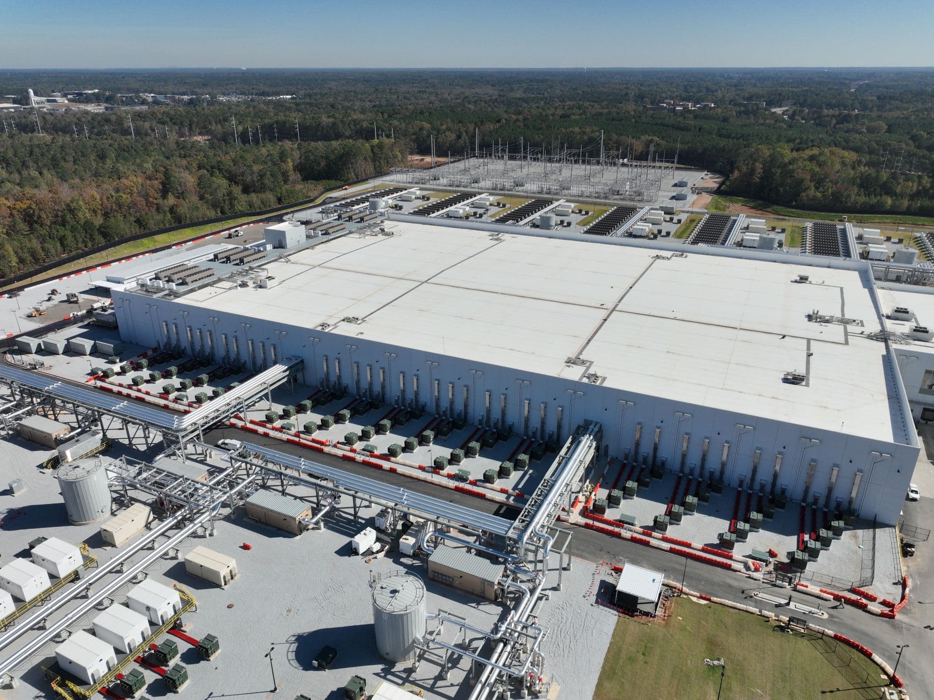 Aerial view of a large gray data center complex with rows of cooling units and white rooftop equipment, surrounded…