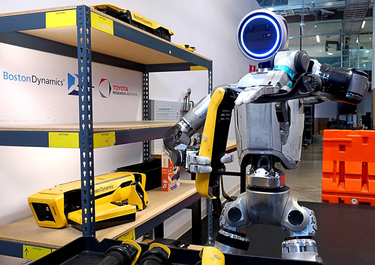 Humanoid robot Atlas hoisting a silver mini-fridge above its waist in a cluttered workshop, with cables and…