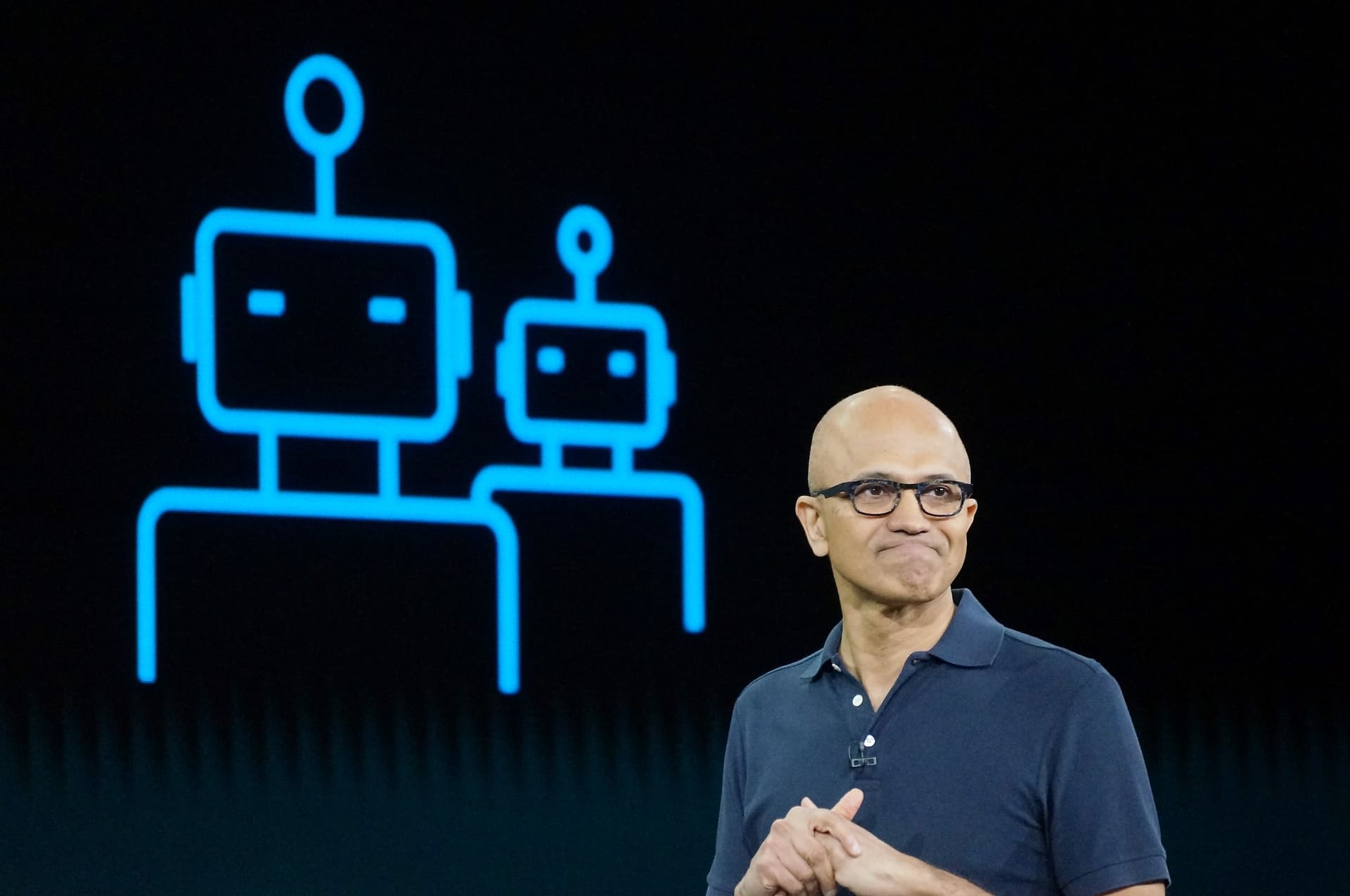 Microsoft CEO Satya Nadella gestures while speaking in an interview, with a blurred background of computer screens…