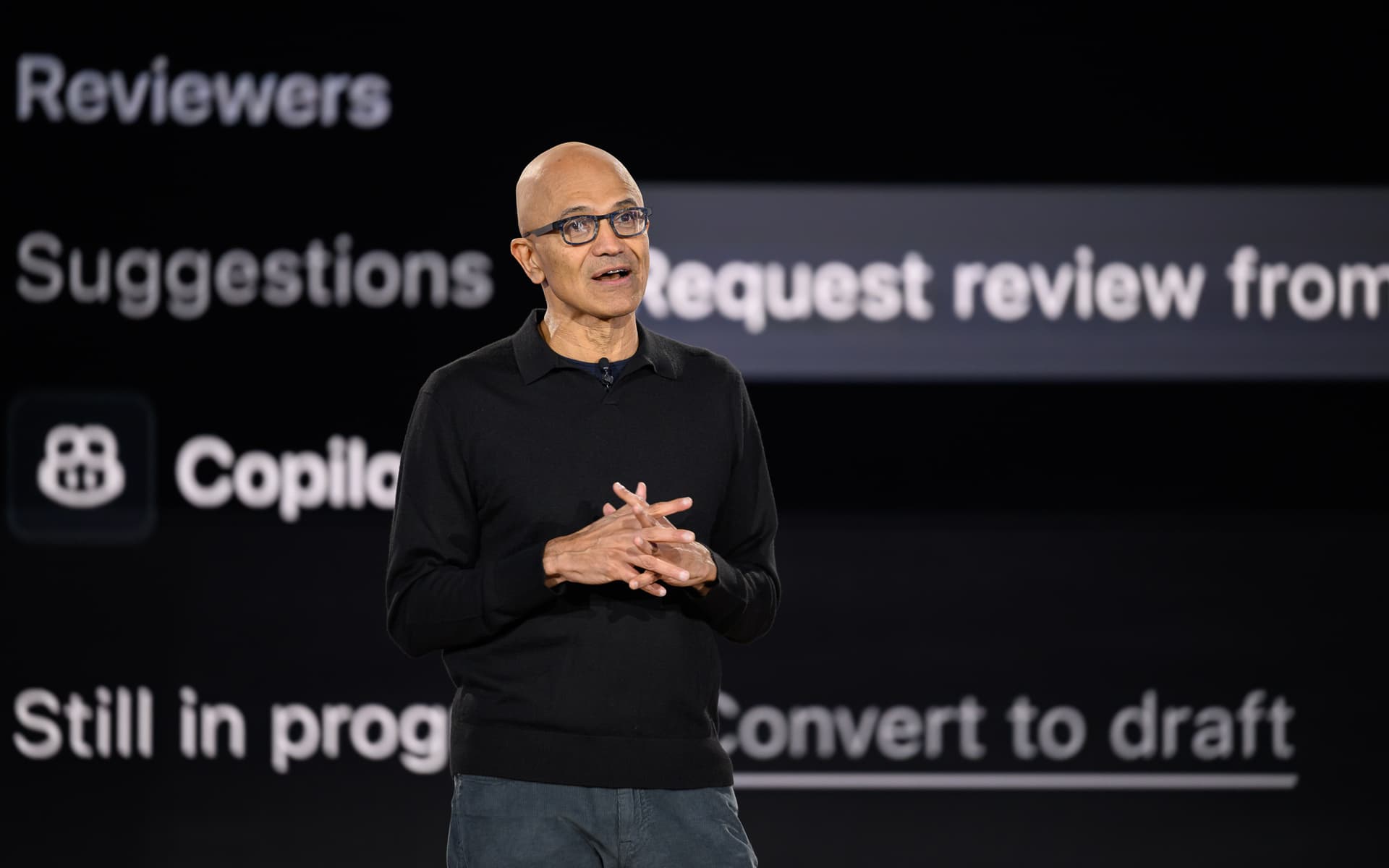 Satya Nadella presenting on stage with a slide showing AI workflow diagrams, addressing an audience in a conference hall