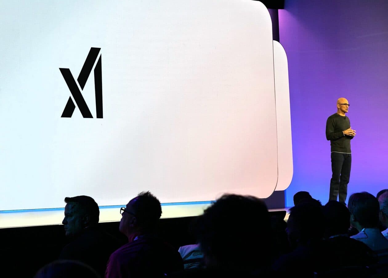 Three tech executives in business attire sit together at a conference table, with logos for Google, Microsoft, and…
