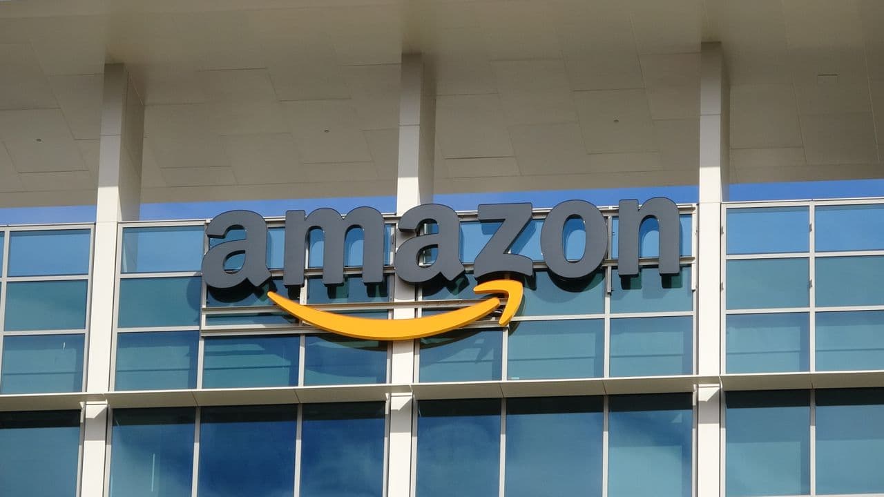 Amazon office workers clustered around monitors, one pointing at a dashboard displaying rising AI token metrics…