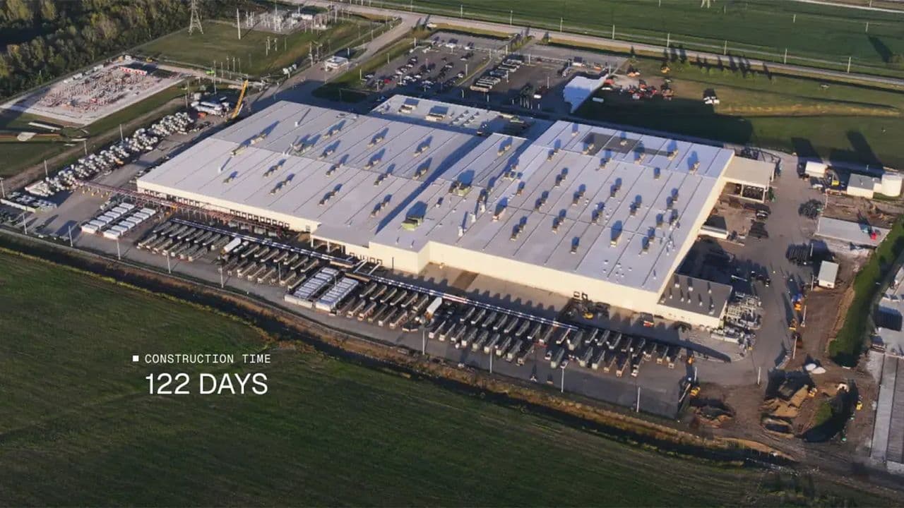 A vast data center interior with rows of server racks and cooling pipes, likely the Colossus 1 supercomputer, leased…
