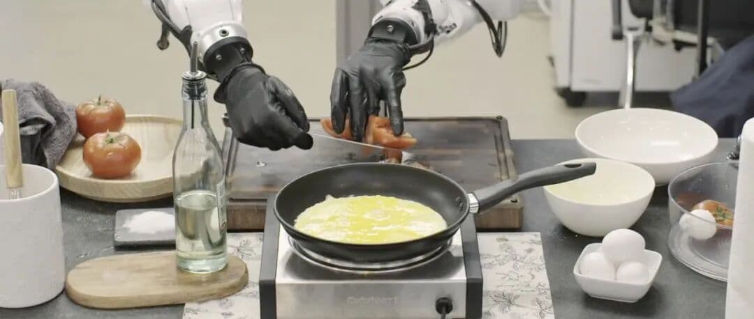 A humanoid robot with a white torso and black arms stands at a kitchen counter, using a spatula to stir-fry tomatoes…