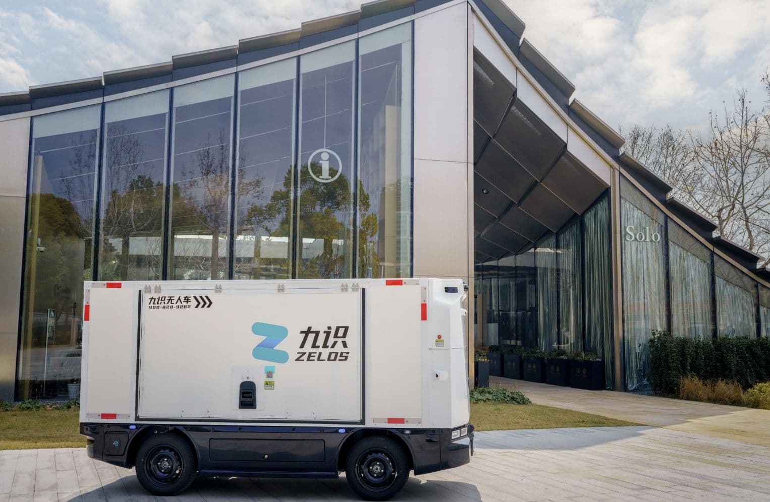 A sleek autonomous electric van with Zelos branding drives through a futuristic logistics hub, surrounded by glowing…