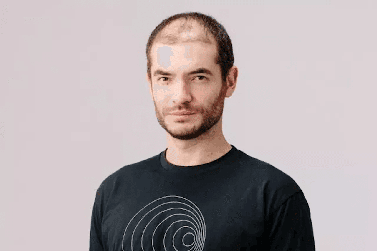 Ilya Sutskever speaks at a tech conference, with a reflective expression, while a blurred OpenAI logo appears in the…