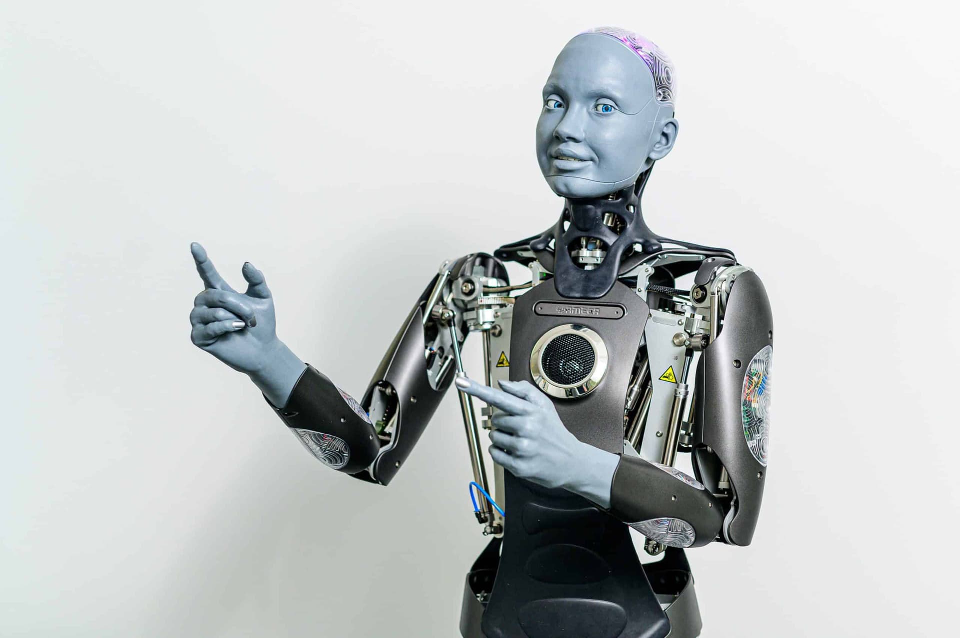 A sleek humanoid robot with a neutral face stands beside a formally dressed First Lady, both facing forward in a…
