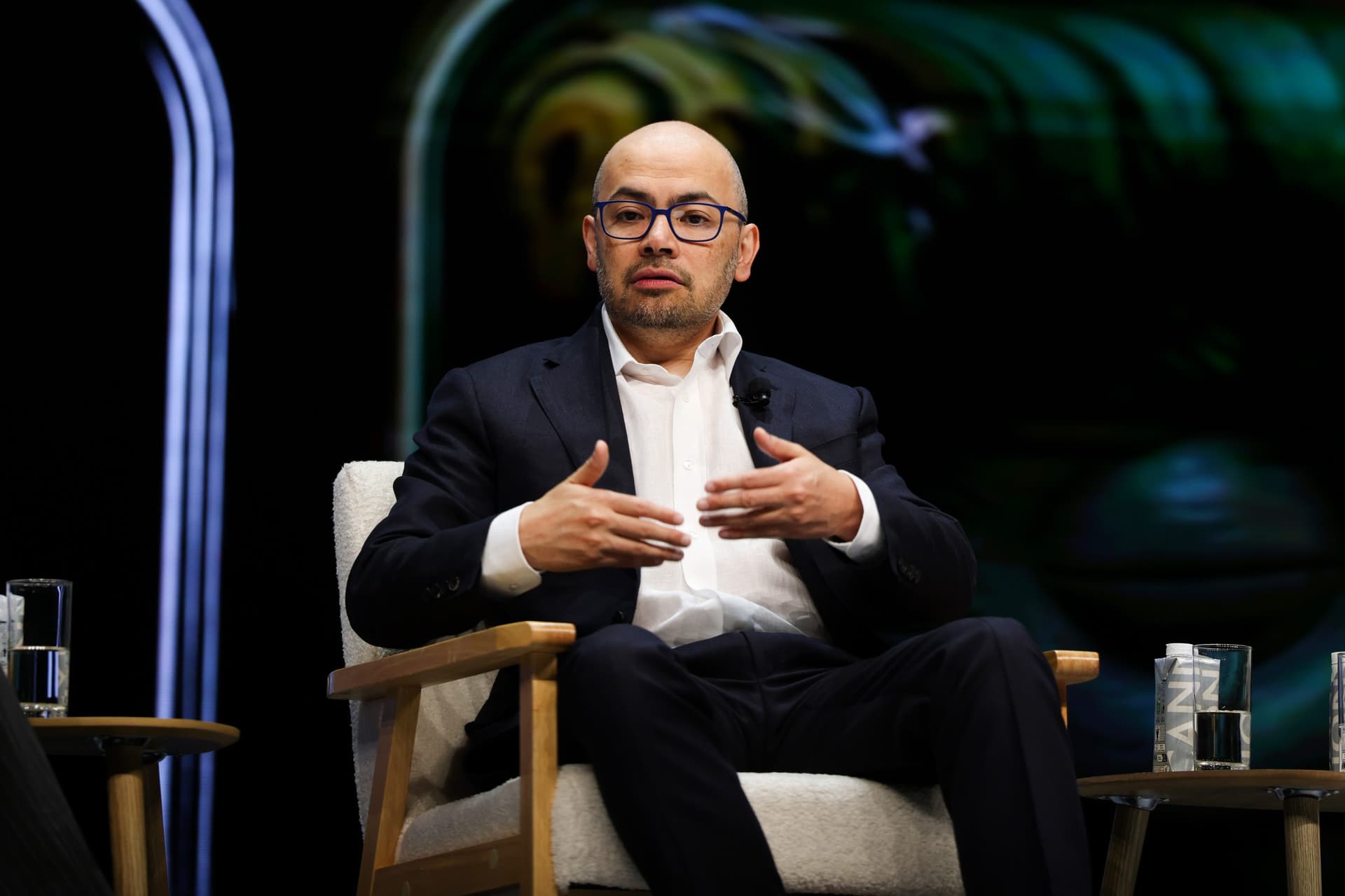 Demis Hassabis at a tech conference, gesturing while speaking on stage, with a DeepMind logo visible behind him