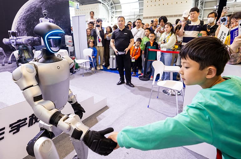 Unitree Robotics humanoid robot standing on a sleek exhibition floor, with engineers adjusting its arm joints under…