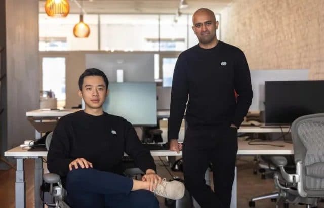 VC George Pu gestures thoughtfully while speaking, with a laptop displaying AI code and a startup pitch deck visible…
