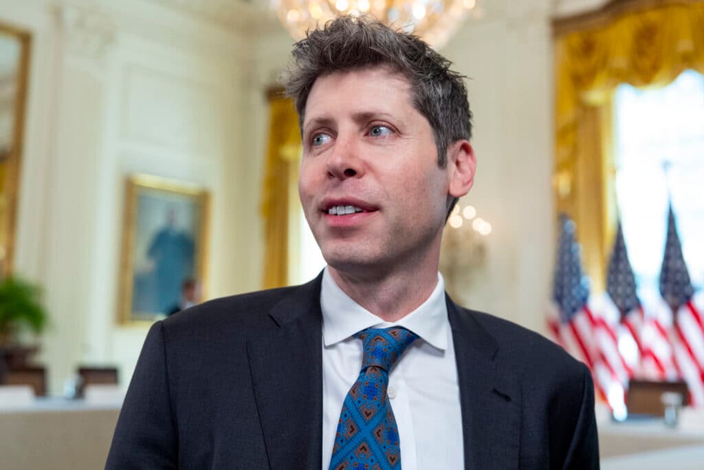Sam Altman speaking at a tech event, with OpenAI logo in background, amid financial pressures and IPO speculation