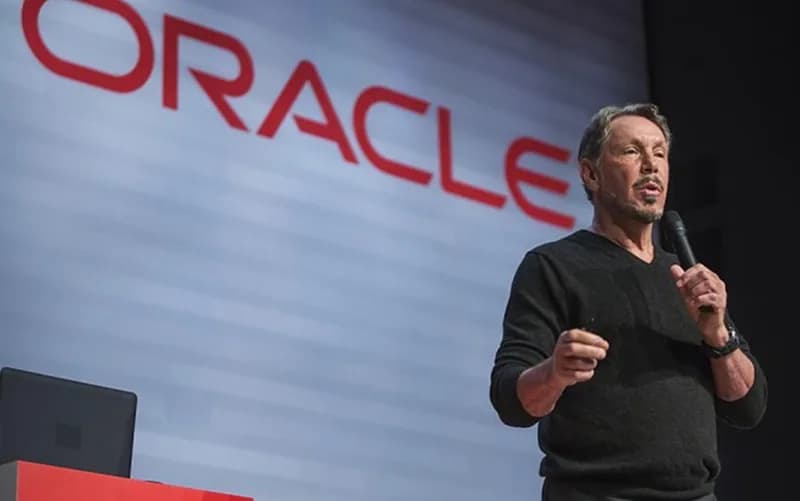 Larry Ellison speaking at a tech conference, with Oracle branding and AI code generation graphics visible on a large…