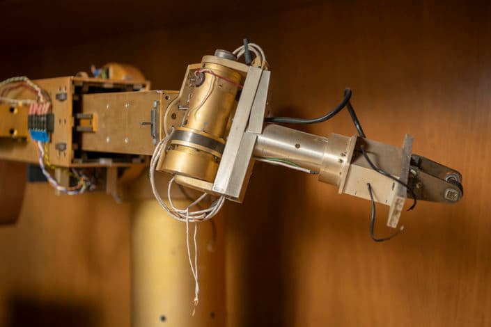 A robotic arm with a gripper holds an object on a lab table, surrounded by electronic equipment and cables