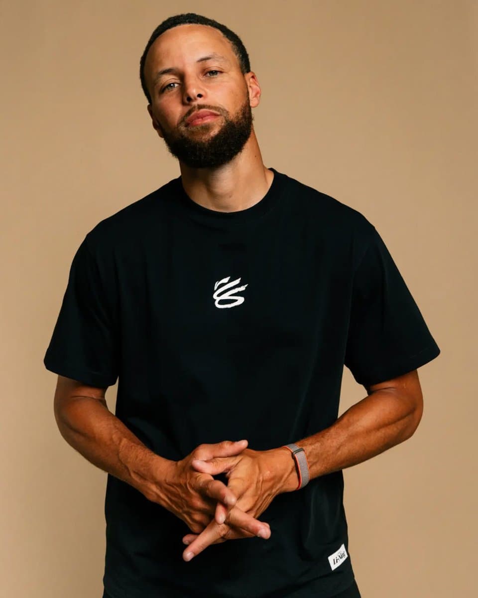 Basketball player Steph Curry holding up a new Li-Ning sneaker with his personal logo visible, standing on a court…