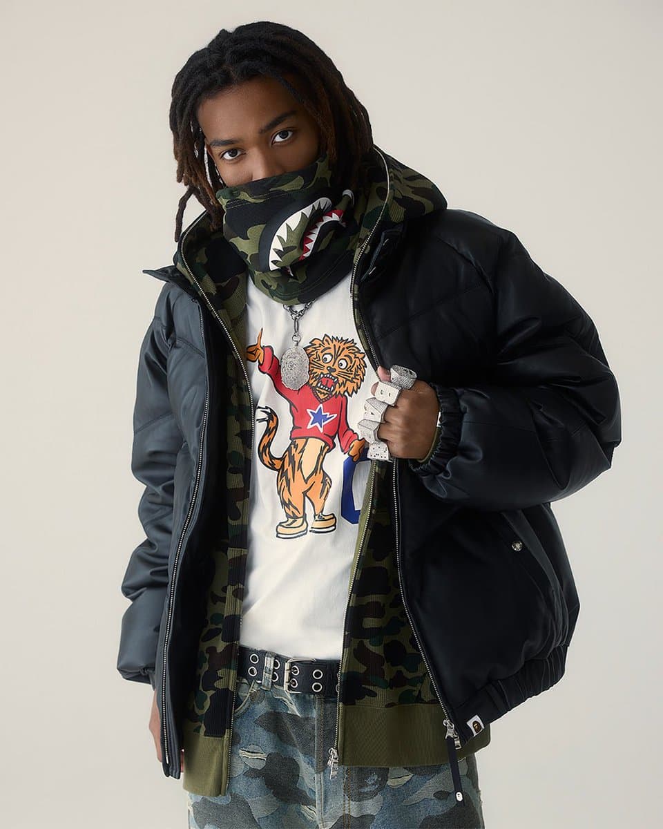 A person wearing a BAPE hoodie and camouflage pants standing in a graffiti-covered alley, holding a skateboard