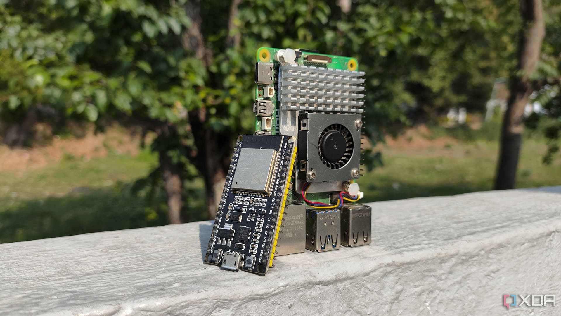 A Raspberry Pi board and an ESP32 microcontroller placed side by side on a desk, with a laptop screen displaying a…