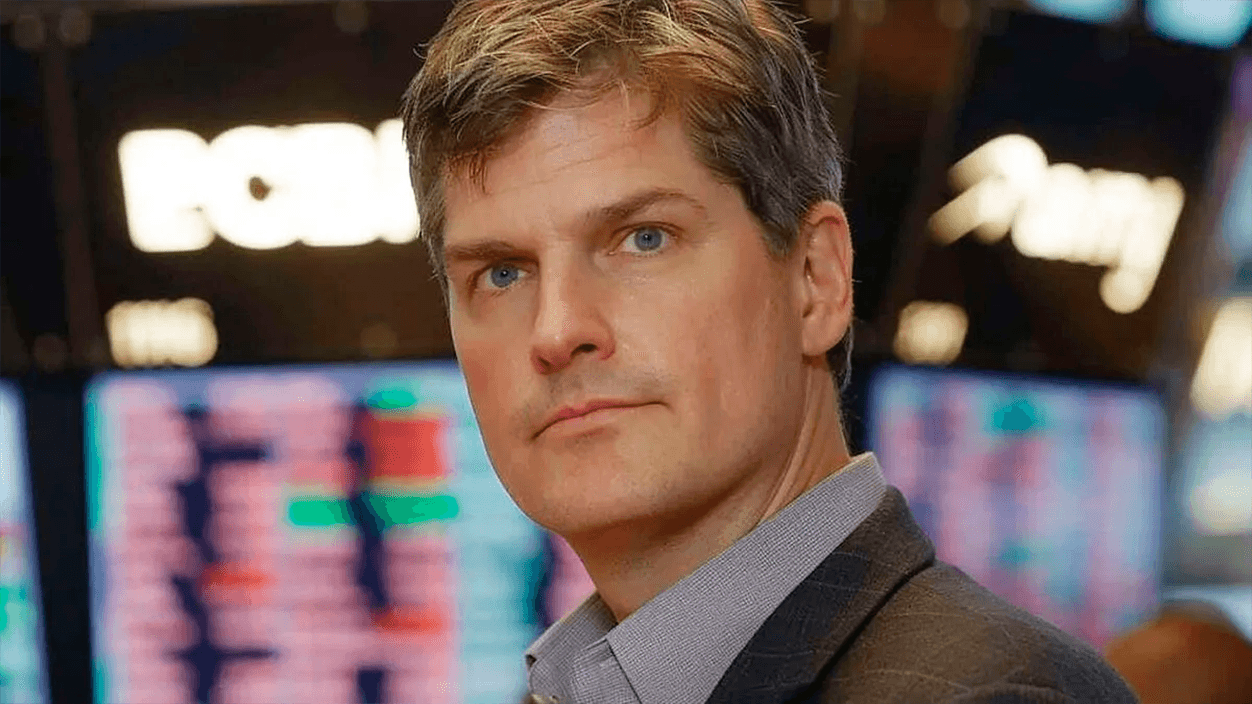 Michael Burry in a suit, pointing at a glowing Anthropic logo on a screen, with Palantir's logo fading in background