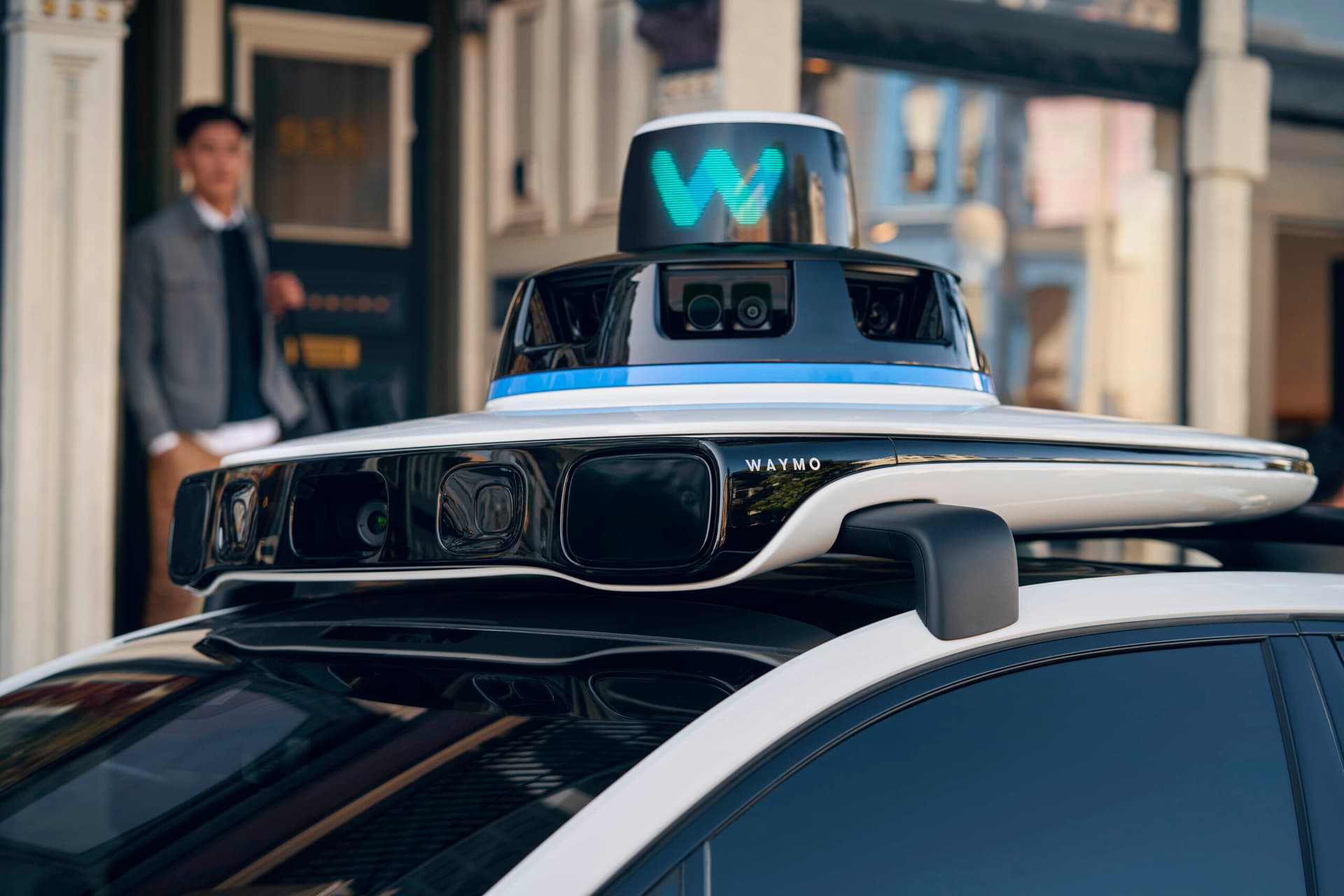 A sleek white Waymo robotaxi with sensor dome parked on a sunlit city street, surrounded by modern buildings and…