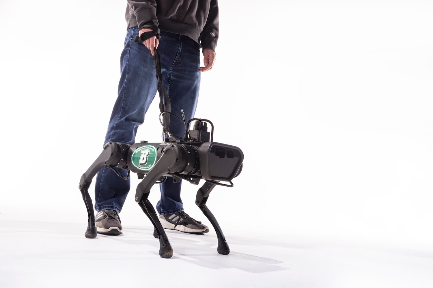 A robotic dog with a leash stands in a lab, a researcher gestures while speaking to it, surrounded by computers and…