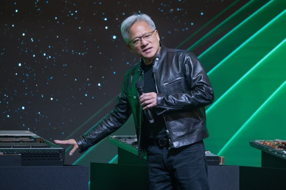 NVIDIA CEO Jensen Huang speaking at a tech conference, gesturing with his hand while standing on stage in front of a…