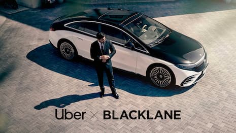 A sleek black chauffeur-driven sedan waits outside a modern glass building while a business traveler in a suit…