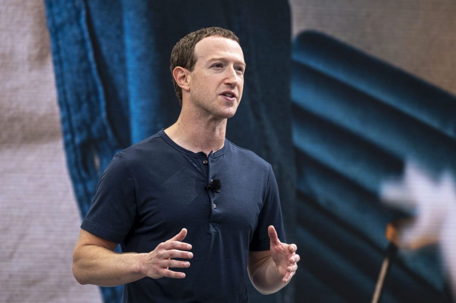 Mark Zuckerberg in casual attire speaking at a tech conference, gesturing while addressing an audience, with a…
