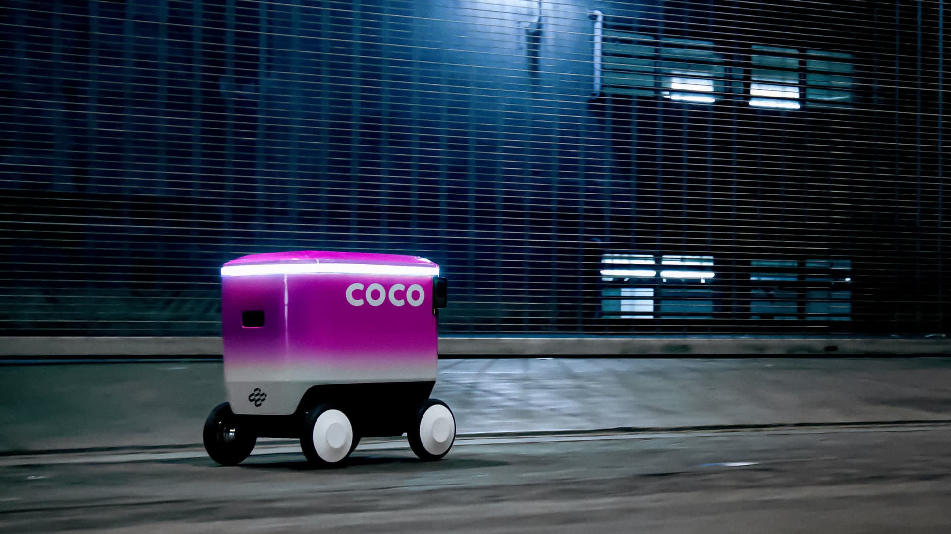 A delivery robot navigates a busy city sidewalk while a person in the background plays Pokémon Go on a smartphone…