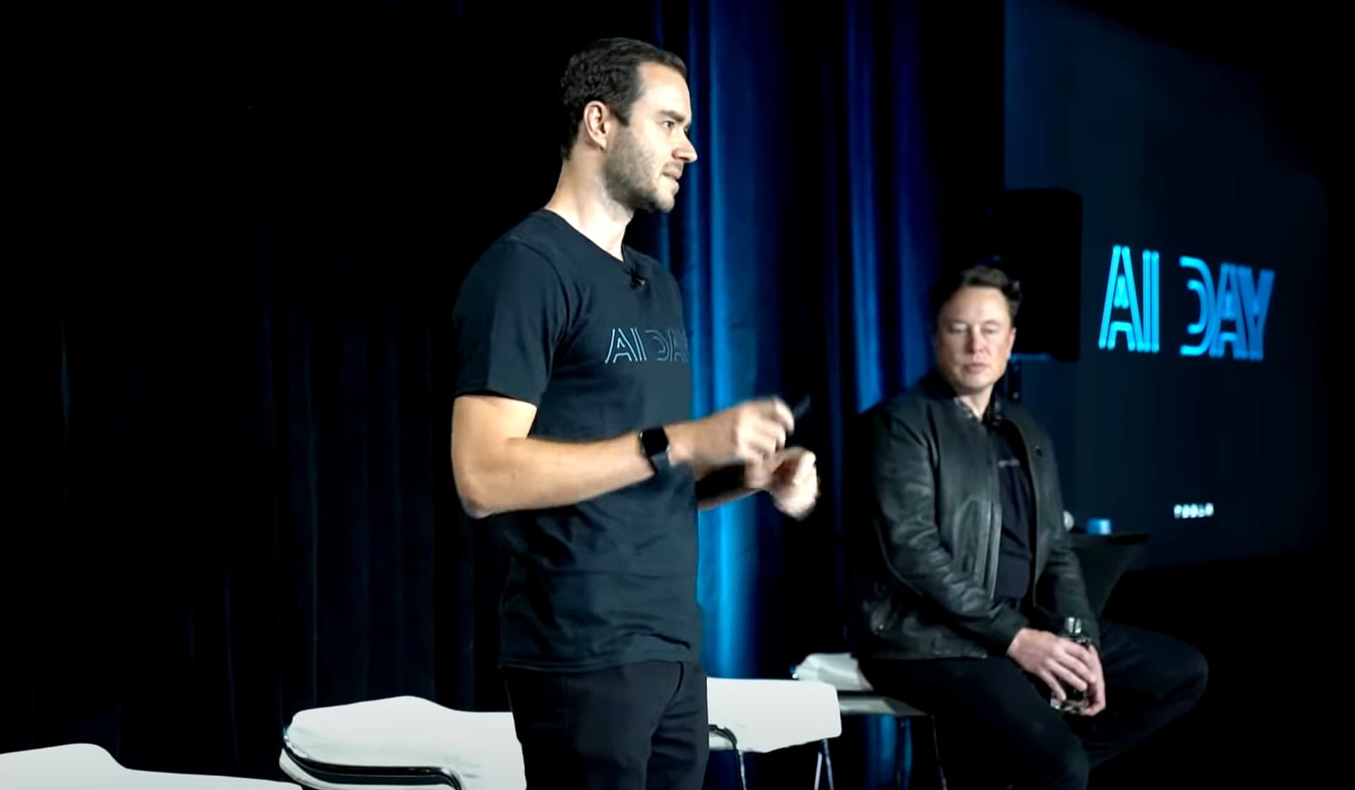 Andrej Karpathy speaking at a tech event, gesturing while discussing AI agent performance and user instruction quality