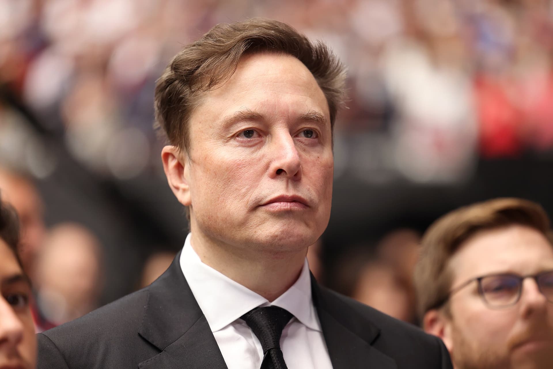 Elon Musk speaking at a podium, likely during a tech conference, with a backdrop of digital screens displaying video…