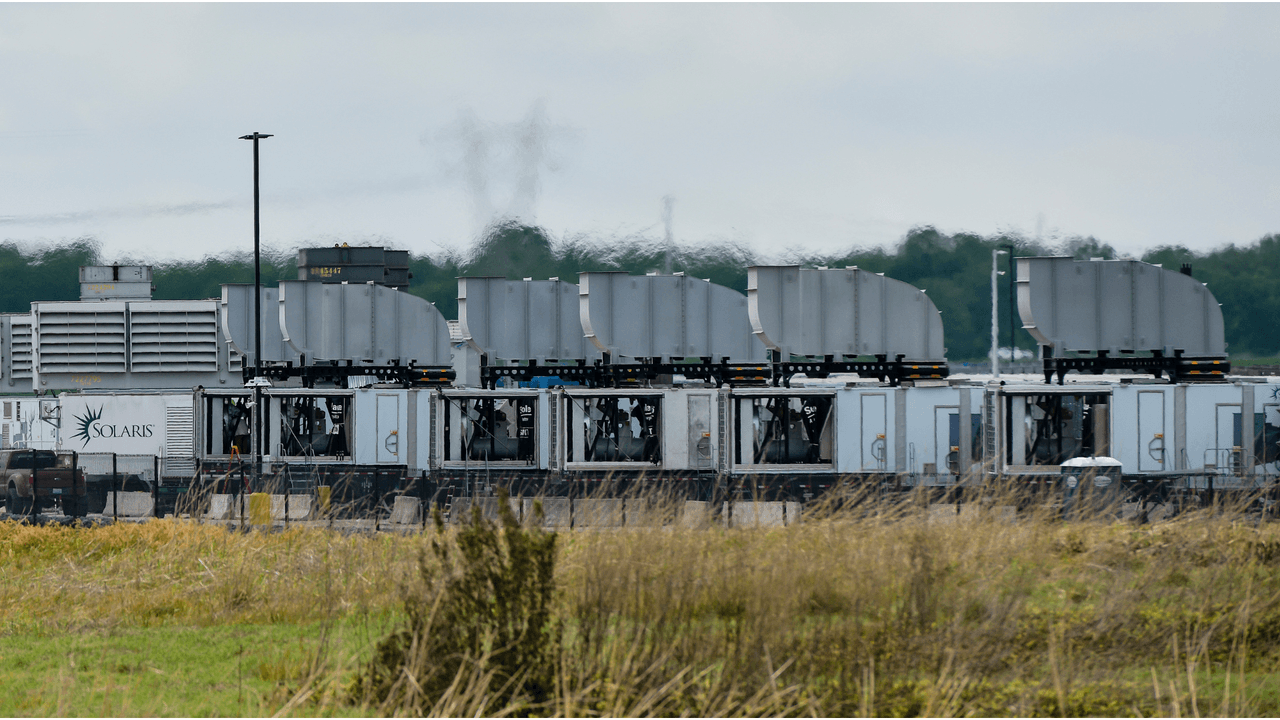 Elon Musk stands beside a large mobile natural gas turbine generator, likely APR Energy equipment, in an industrial…