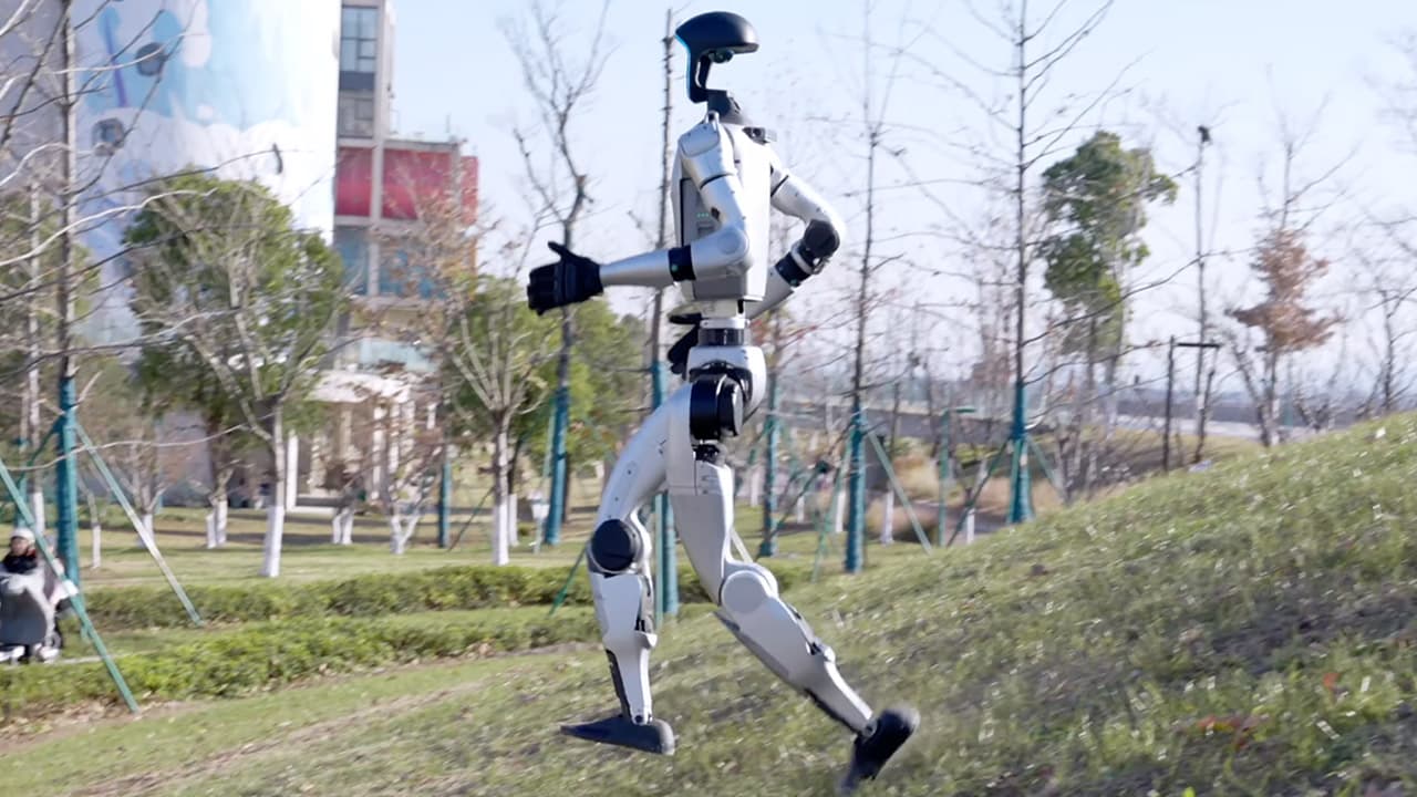 A sleek white humanoid robot stands upright indoors, arms at its sides, amidst a retail or event setting with…