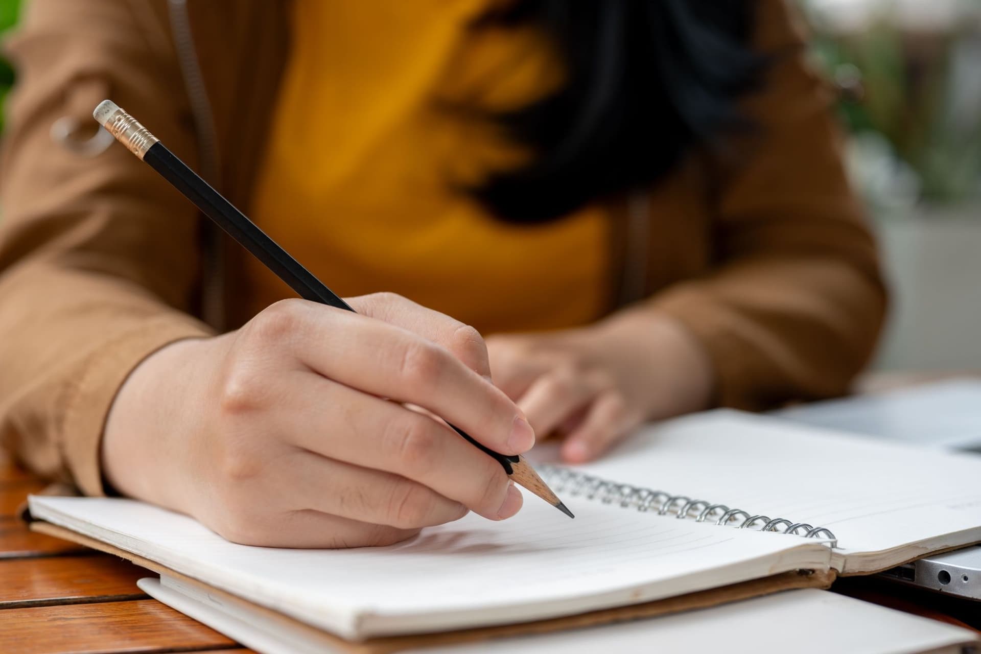 A woman holds a pencil and writes in a notebook at a desk, reflecting a return to handwritten assignments in…